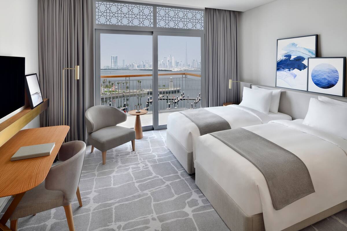 A hotel room with a large bed, a desk with a chair, a small table, and a view of the city through large windows.