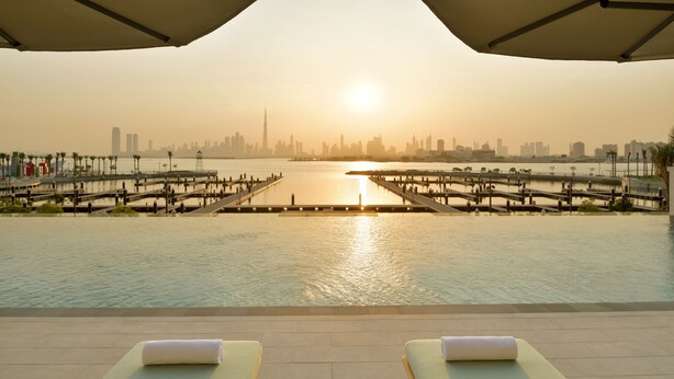 A waterfront view with a city skyline in the background, a sunlit pool area with lounge chairs, and a bridge connecting two sides of the pool. - Vida Creek Harbour (Dubai)