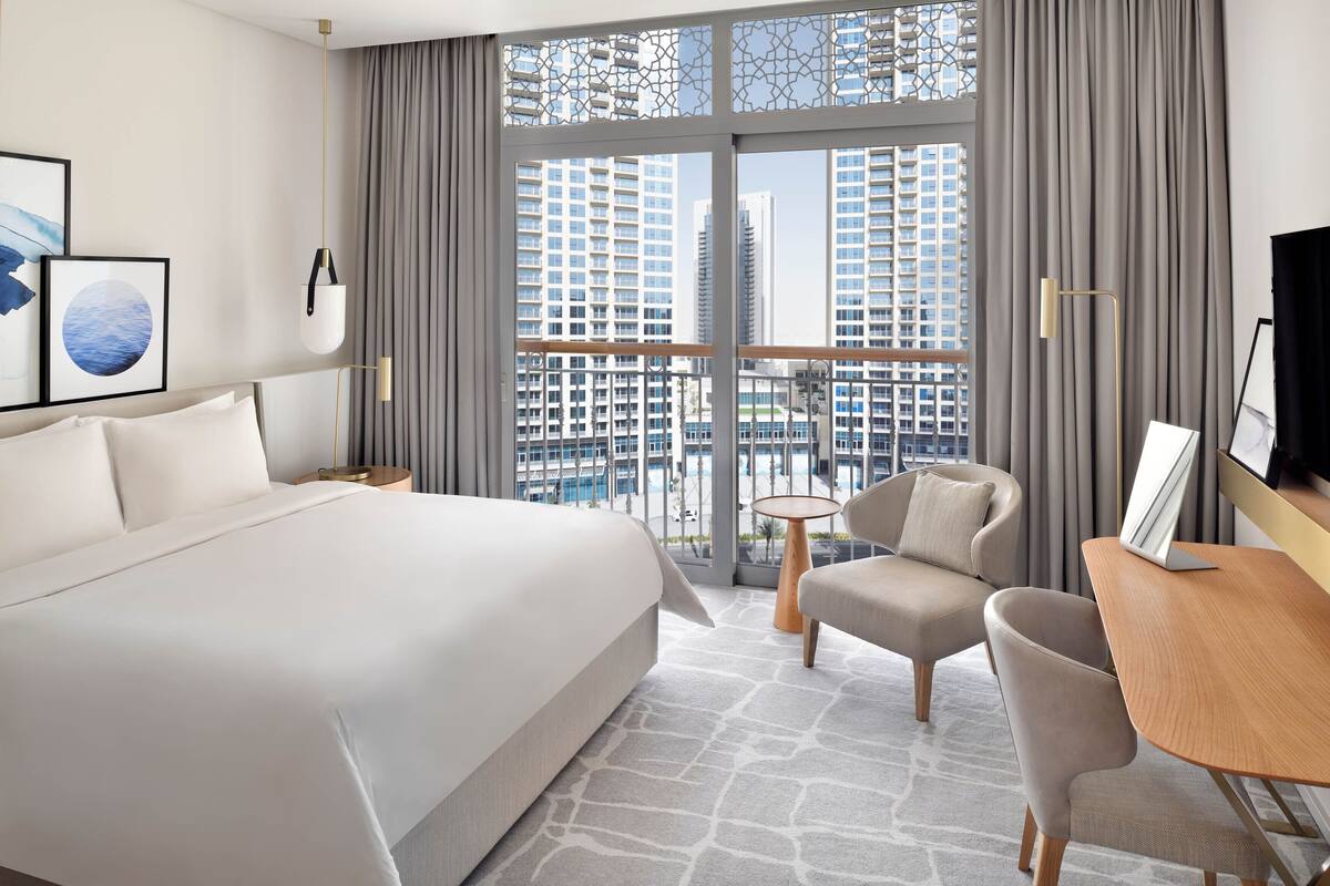 A hotel room with a large bed, a desk, a chair, and a view of tall buildings through large windows.
