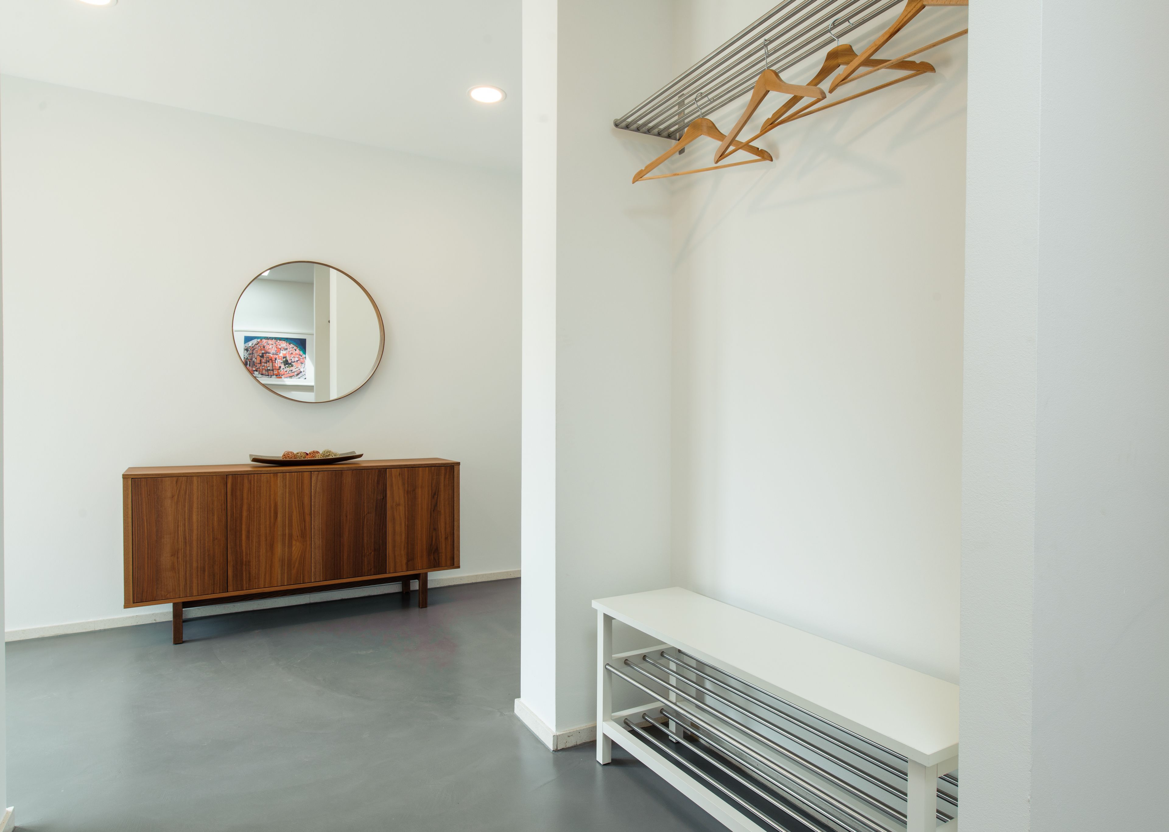 A modern, minimalist room with a wooden cabinet, a round mirror, and a white bench.