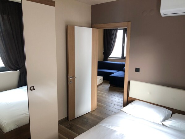 Habitación de hotel con piso de madera, una cama con ropa blanca, un sofá y una ventana con cortinas.