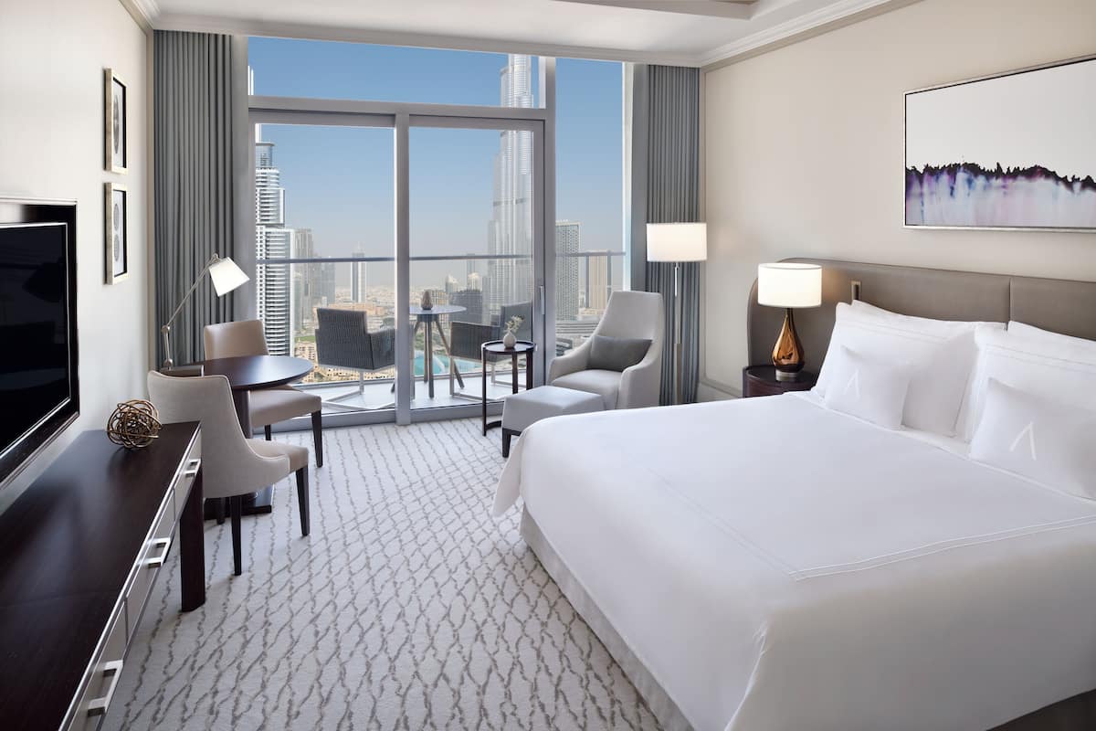A hotel room with a large bed, a desk, a chair, a small table, and a TV. The room has a view of a cityscape with tall buildings.