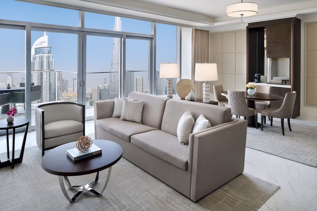 A modern hotel room with a large window offering a city view, a sofa, a coffee table, and a dining area with chairs.