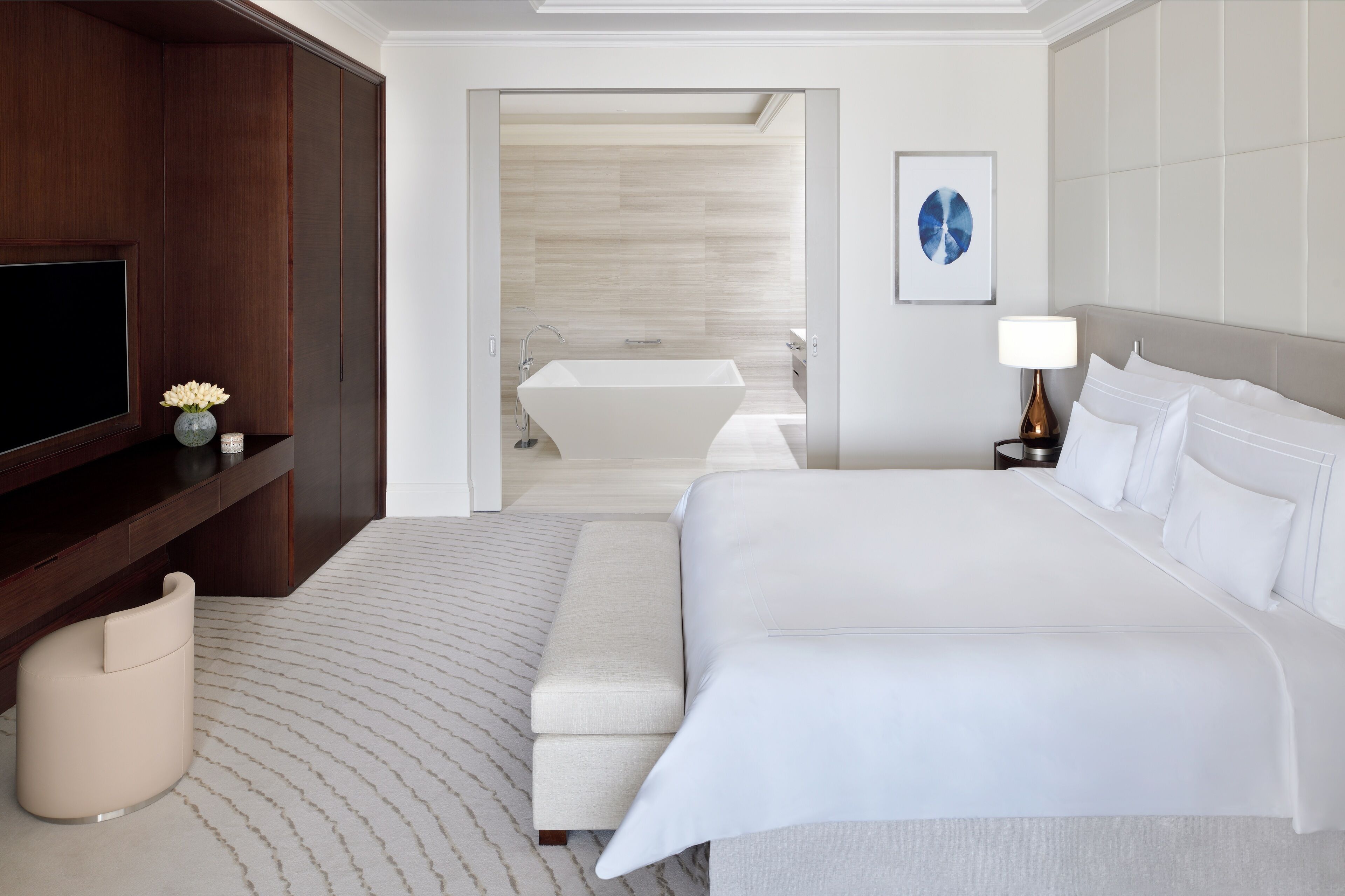 A modern hotel room with a large bed, a flat-screen TV, a bedside table with a lamp, and a view into the bathroom.