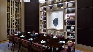 A dining area with a dark wooden table set for four, surrounded by matching chairs. A large bookshelf with various decorative items and a framed artwork is behind the table.