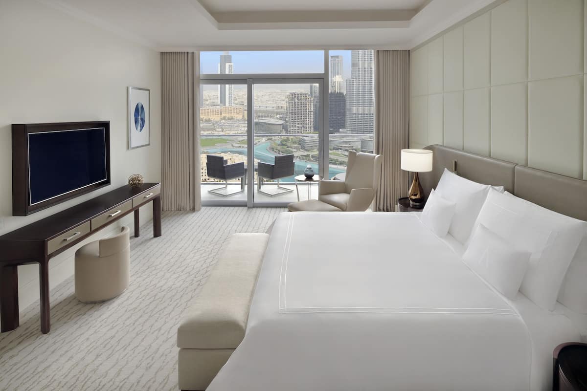 A hotel room with a large bed, a desk with a TV, a chair, a lamp, and a view of the cityscape through large windows.