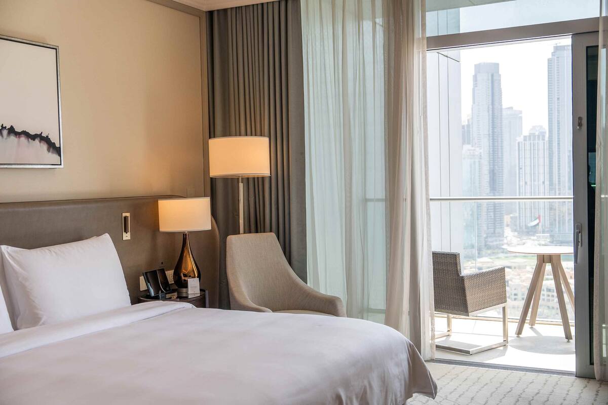 A hotel room with a large bed, a chair, a desk with a lamp, and a view of a cityscape through a large window.