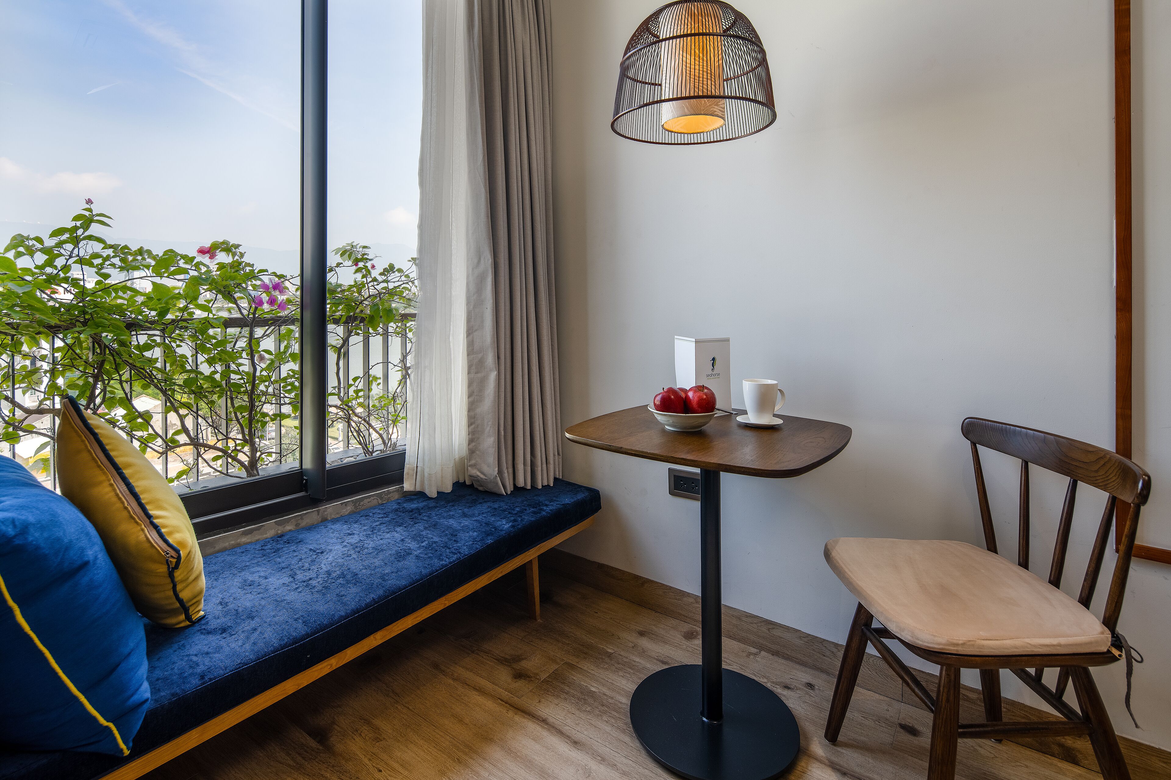 A balcony with a blue cushioned bench, a wooden chair, a small round table with a bowl of fruit, a white vase, and a hanging light fixture.