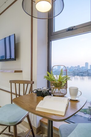 A wooden table with a book, a camera, and a plant. A television is mounted on the wall, and a window offers a view of a city skyline. - Seahorse Han Market Da Nang Hostel by Haviland (Da Nang)