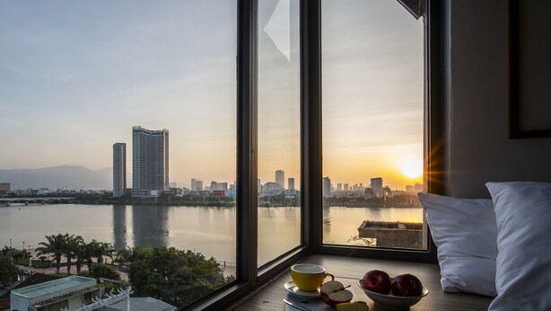 A room with a large window overlooking a cityscape at sunset, a table with apples and a cup, and a view of a river. - Seahorse Han Market Da Nang Hostel by Haviland (Da Nang)