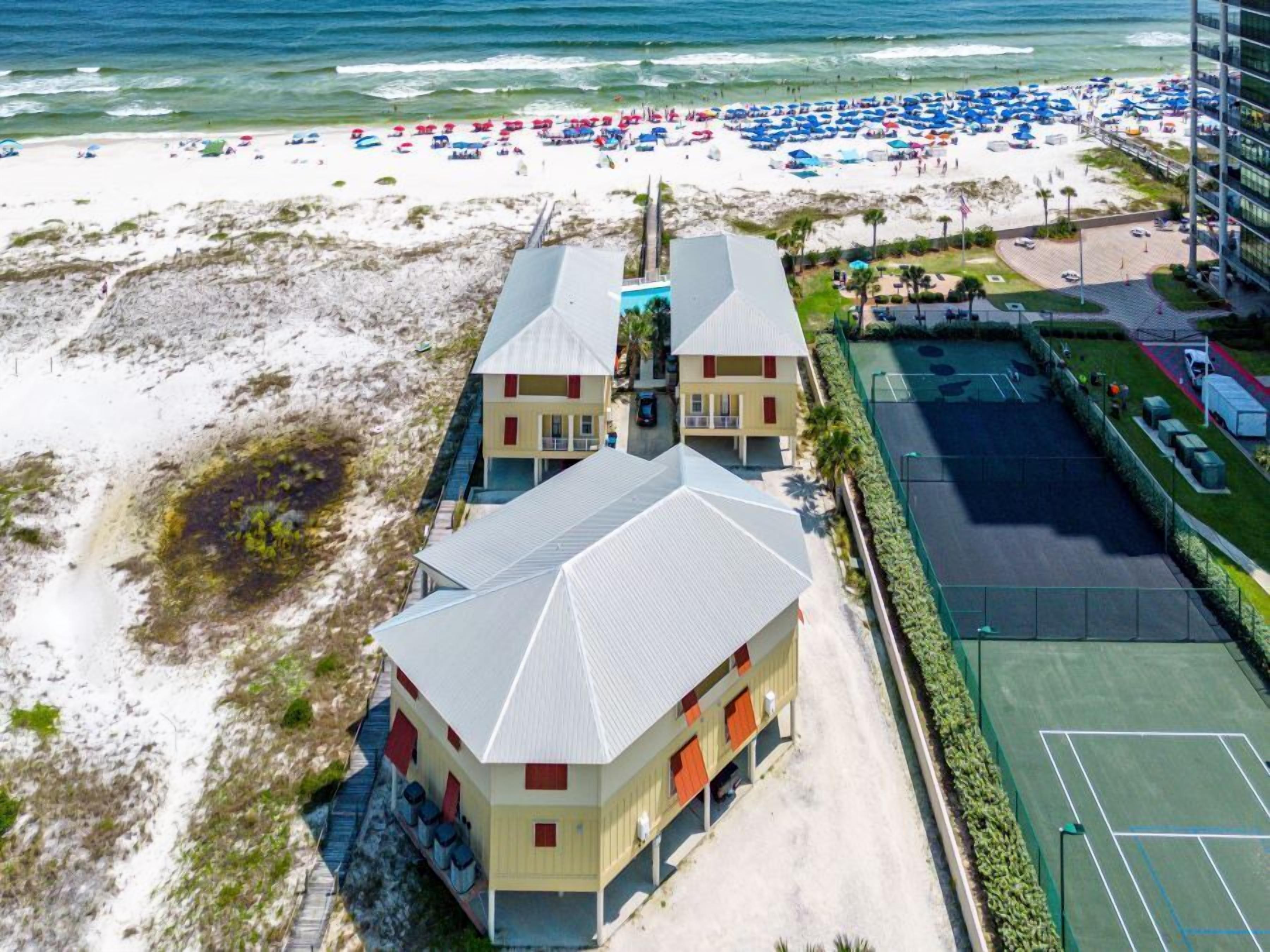 Un hôtel en bord de mer, avec des bâtiments à deux étages, une plage de sable dotée de parasols et un court de tennis.