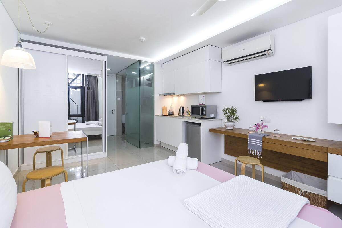 Quarto de hotel moderno com uma cama, uma escrivaninha, uma cadeira, uma TV, um micro-ondas e uma pequena cozinha equipada.