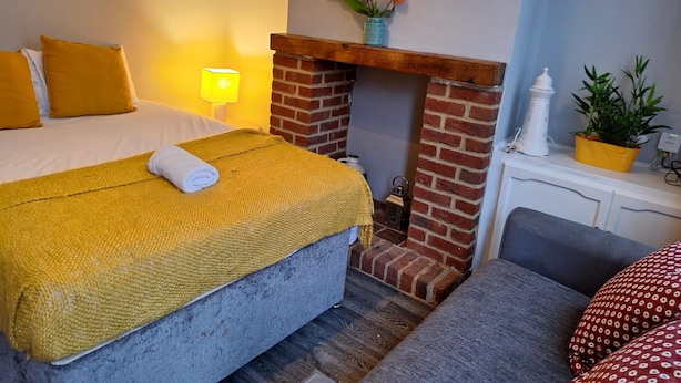 A hotel room with a bed, a small fireplace, a nightstand, a lamp, and a painting on the wall. - Huku Kwetu - Ferndale House (Luton)
