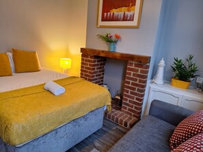 A hotel room with a bed, a small fireplace, a nightstand, a lamp, and a painting on the wall. - Huku Kwetu - Ferndale House (Luton)