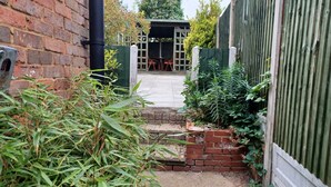 A brick wall, a metal pipe, a green fence, and a pathway leading to a covered area with a table and chairs. - Huku Kwetu - Ferndale House (Luton)