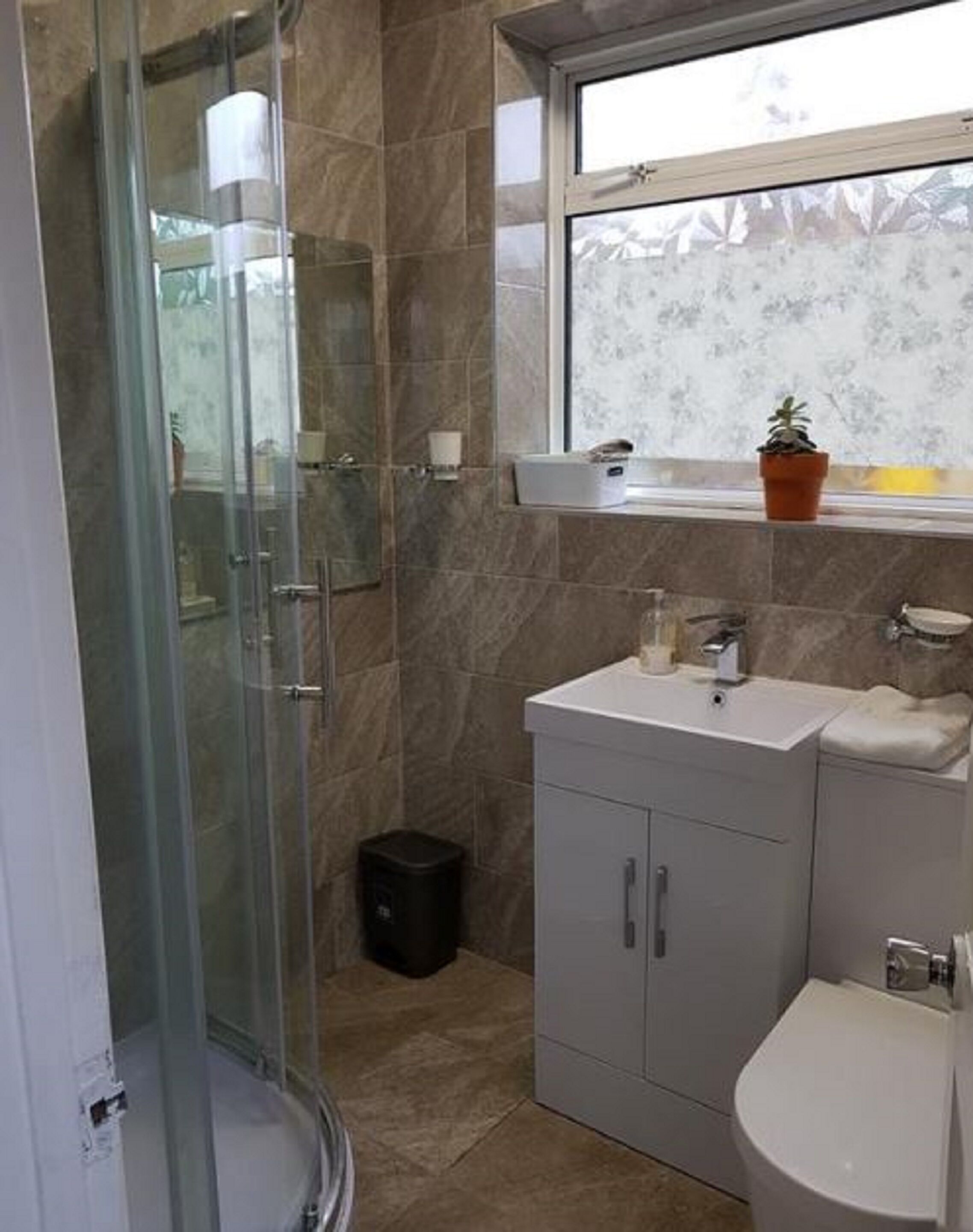 A bathroom with a glass shower enclosure, a sink with a mirror, a window above the sink, and a potted plant on the windowsill.