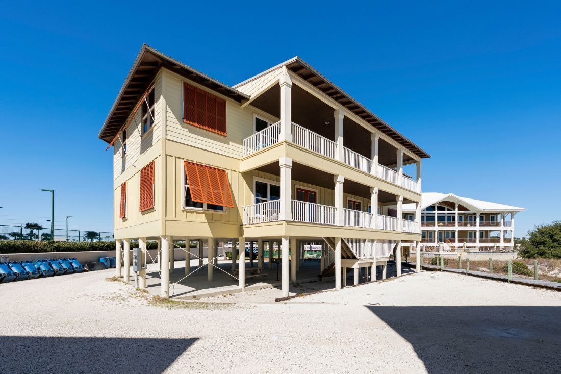 A two-story beachfront house with multiple balconies, a covered porch, and a sandy beach in the background.
