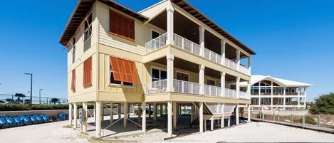 A two-story beachfront house with multiple balconies, a covered porch, and a sandy beach in the background.