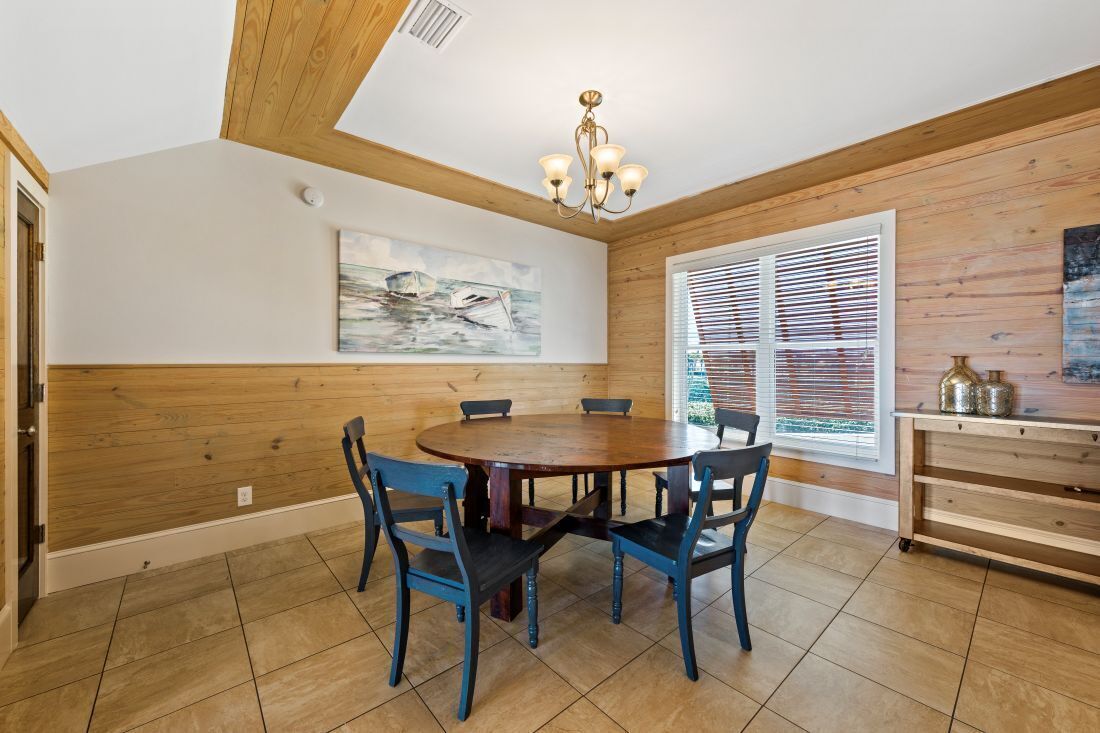 A dining area with a wooden table and chairs, a painting on the wall, and a window with blinds.