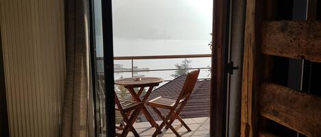 Una habitación con una cama, una silla de madera, una mesita y vistas a una montaña y al agua a través de una puerta abierta.