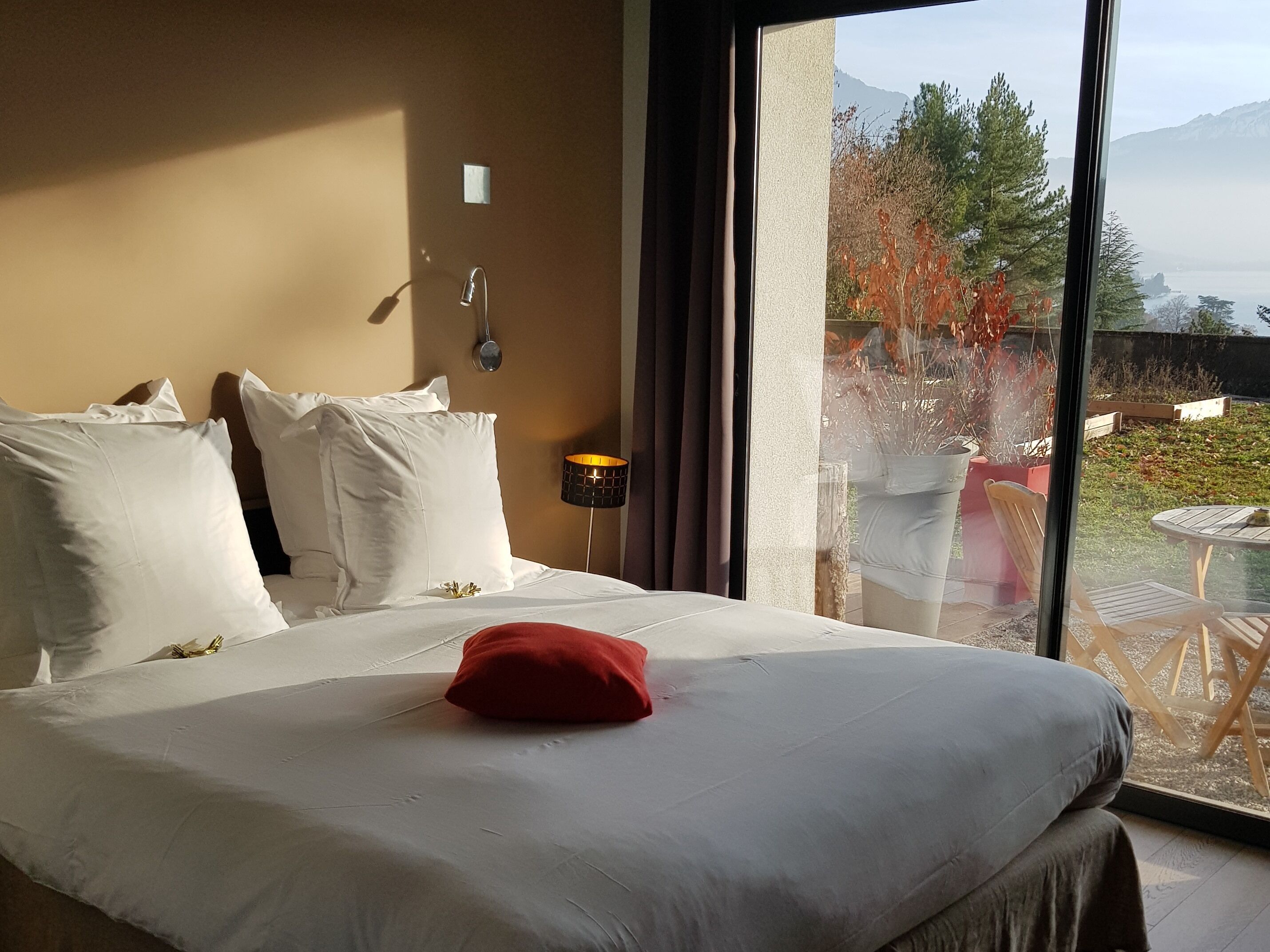Una habitación de hotel con una cama grande, ropa de cama blanca, una almohada roja y vistas al exterior a través de un ventanal.