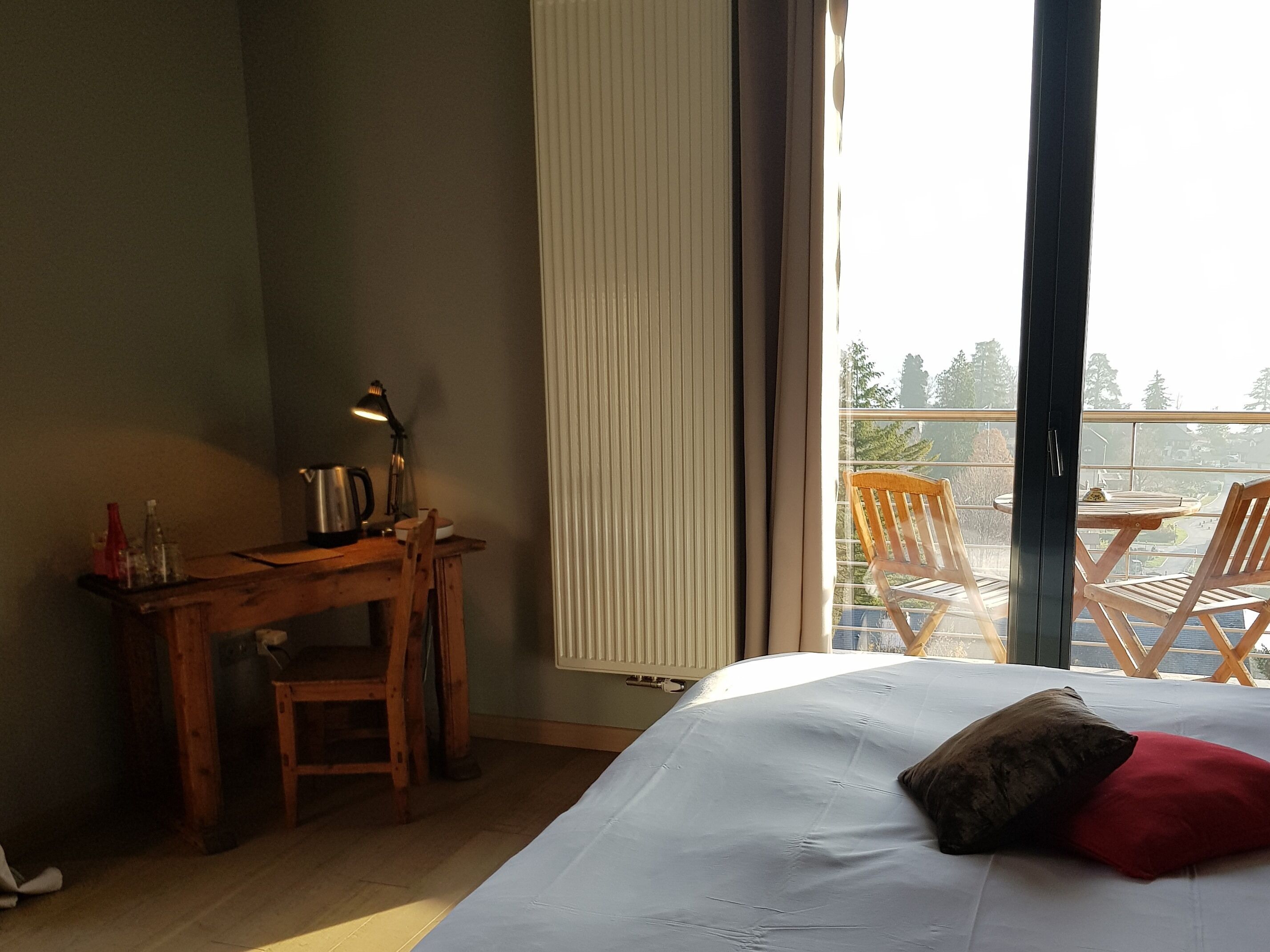 A bedroom with a bed, a desk, and a chair. There is a window with blinds and a view of the outside.