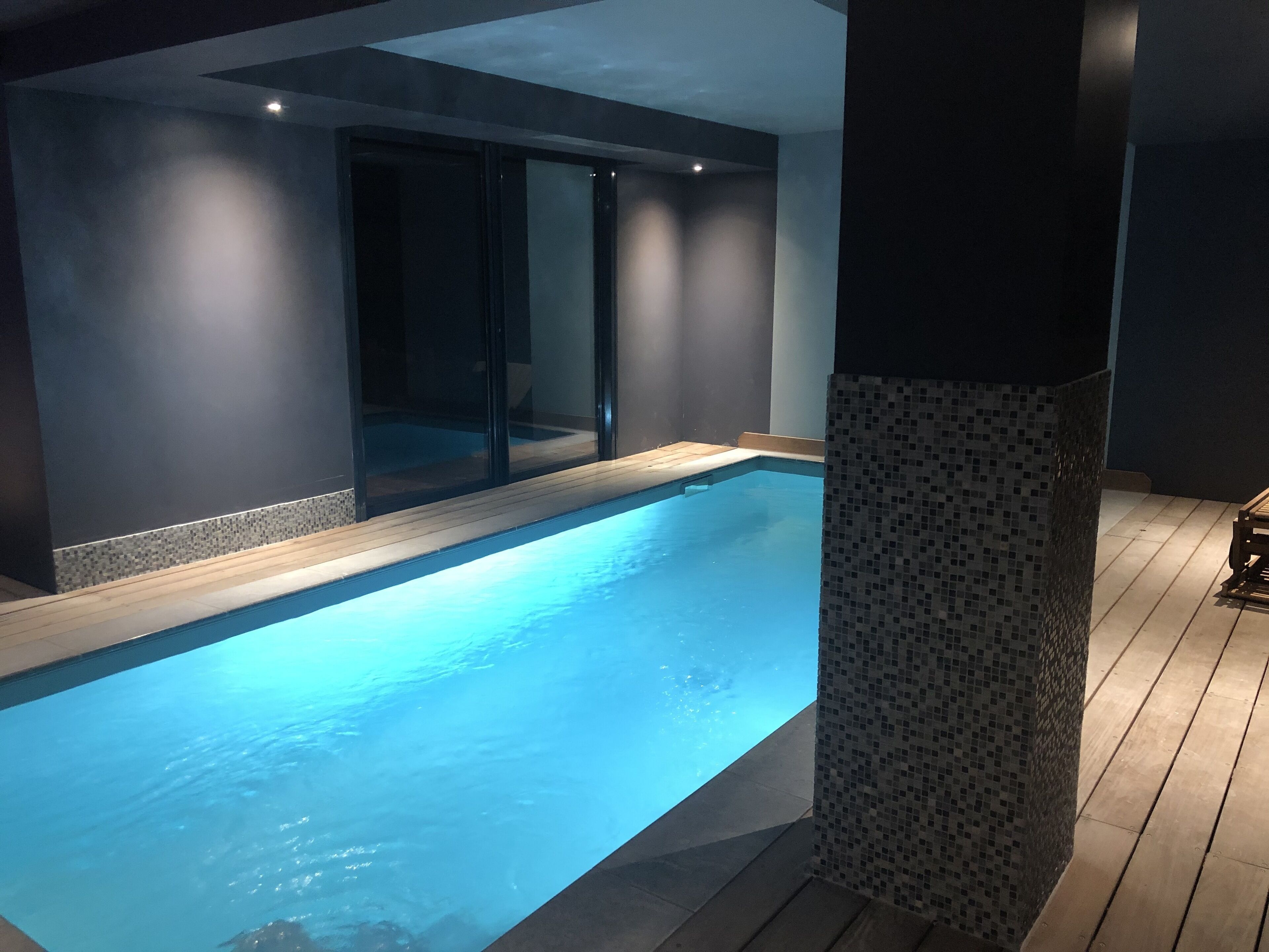 An indoor swimming pool with a tiled edge, a wooden deck, and a mosaic column.