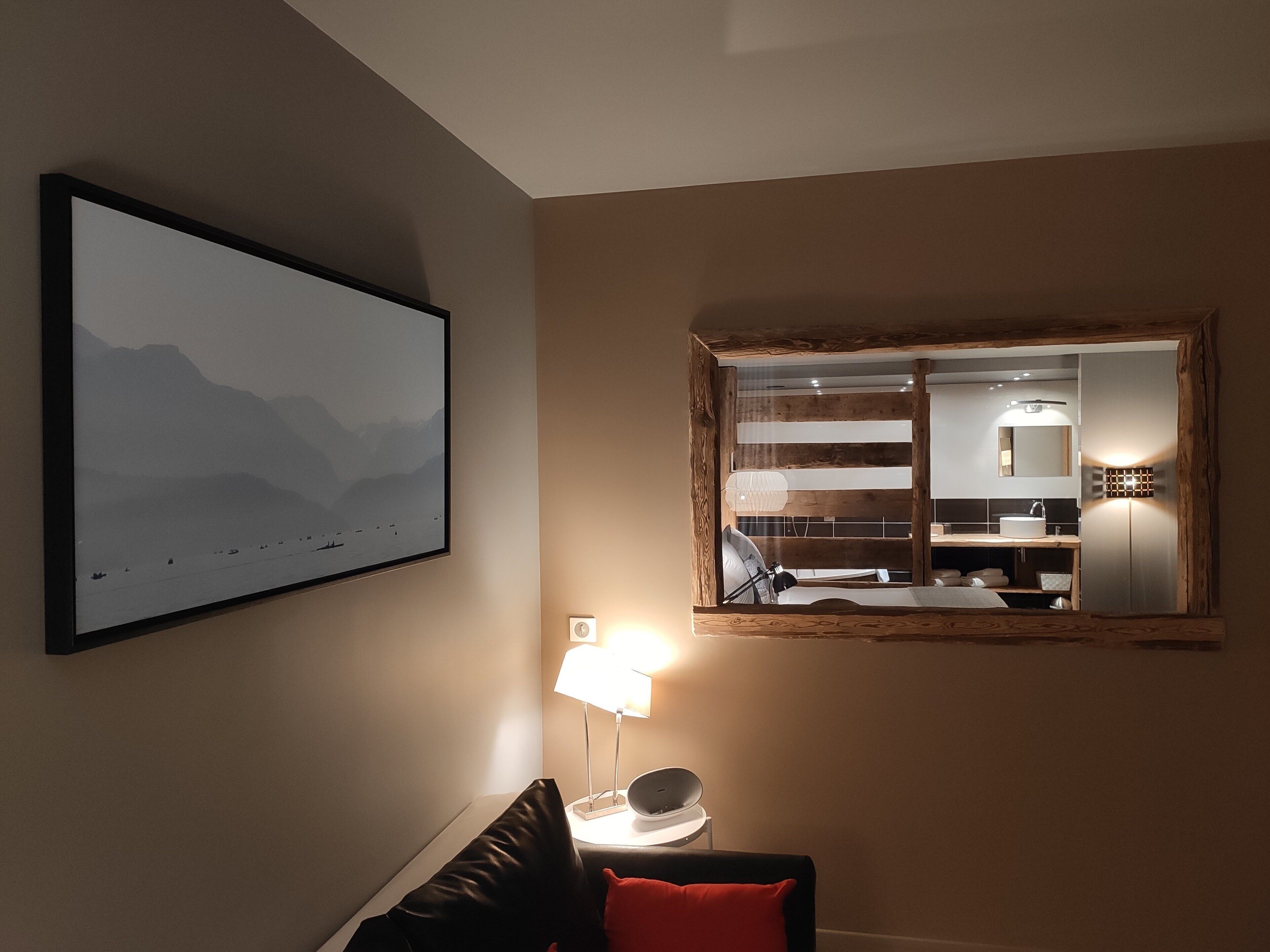 A modern hotel room with a large framed mirror, a mounted picture of mountains, a bedside lamp, and a view of the bathroom.