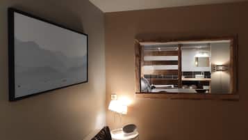 A modern hotel room with a large framed mirror, a mounted picture of mountains, a bedside lamp, and a view of the bathroom.