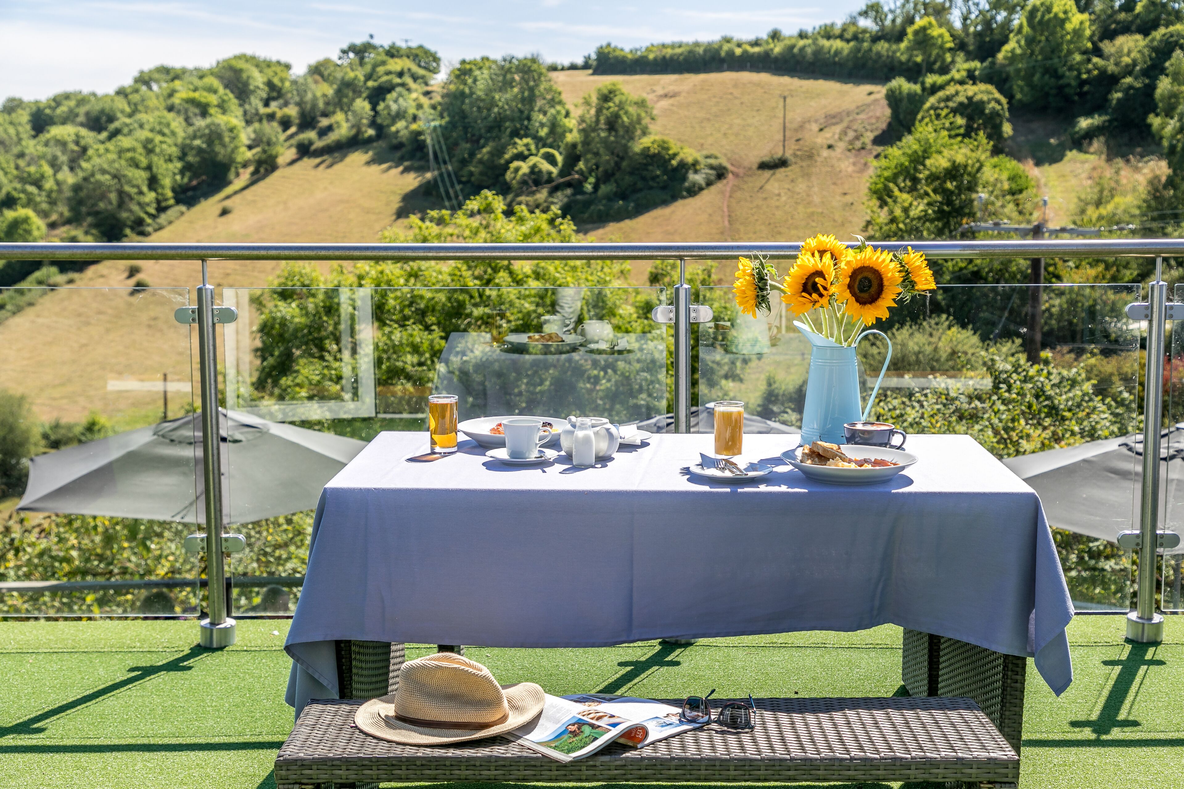A table set with breakfast items, a sunflowers vase, and a hat on a balcony with a scenic view.