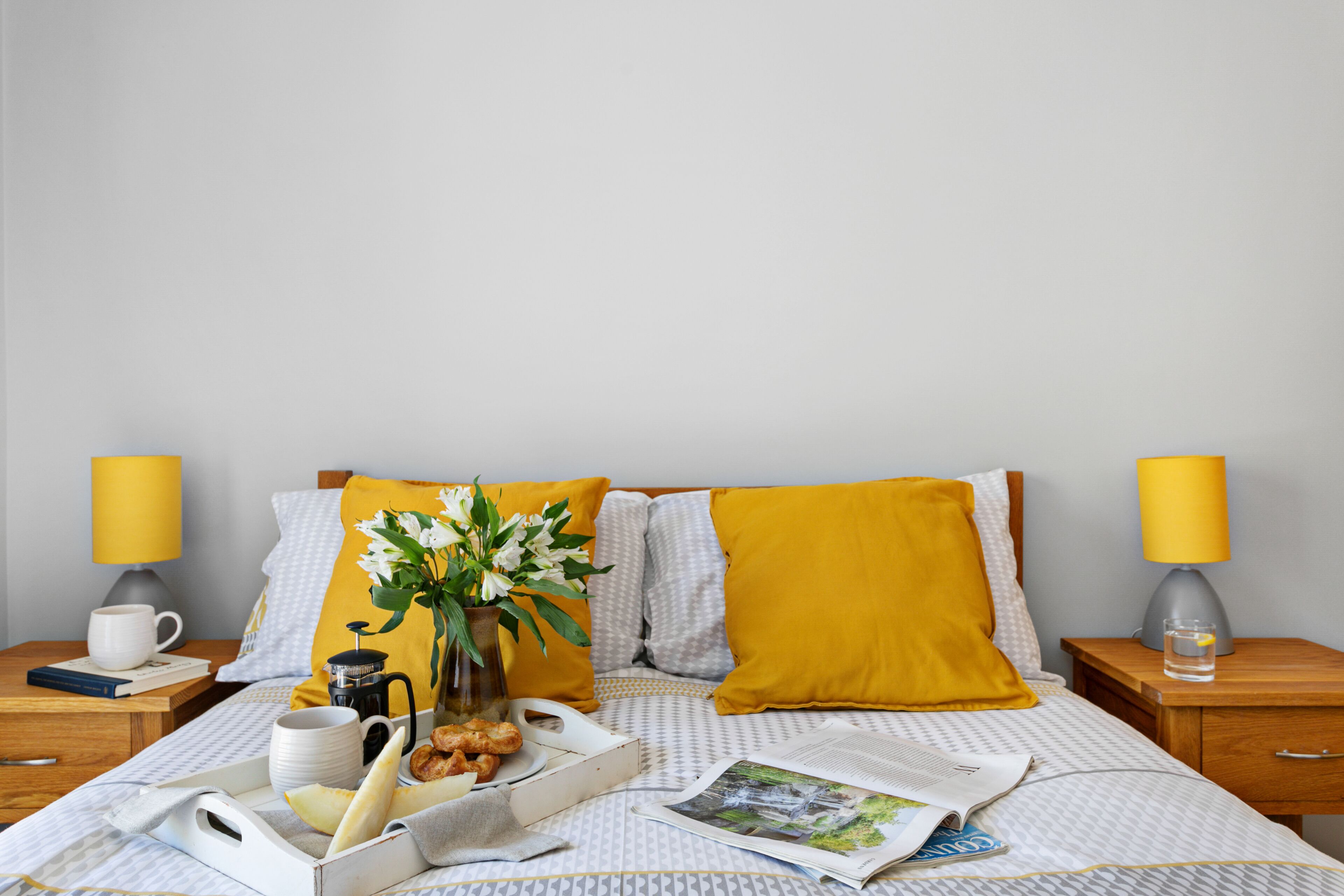 Un letto rifatto con un cuscino giallo, un comodino con una lampada e un vassoio con prodotti per la colazione.