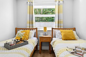 A bedroom with two beds, a bedside table, a window with curtains, and a view of greenery outside. - Old Walls Vineyard (Teignmouth)