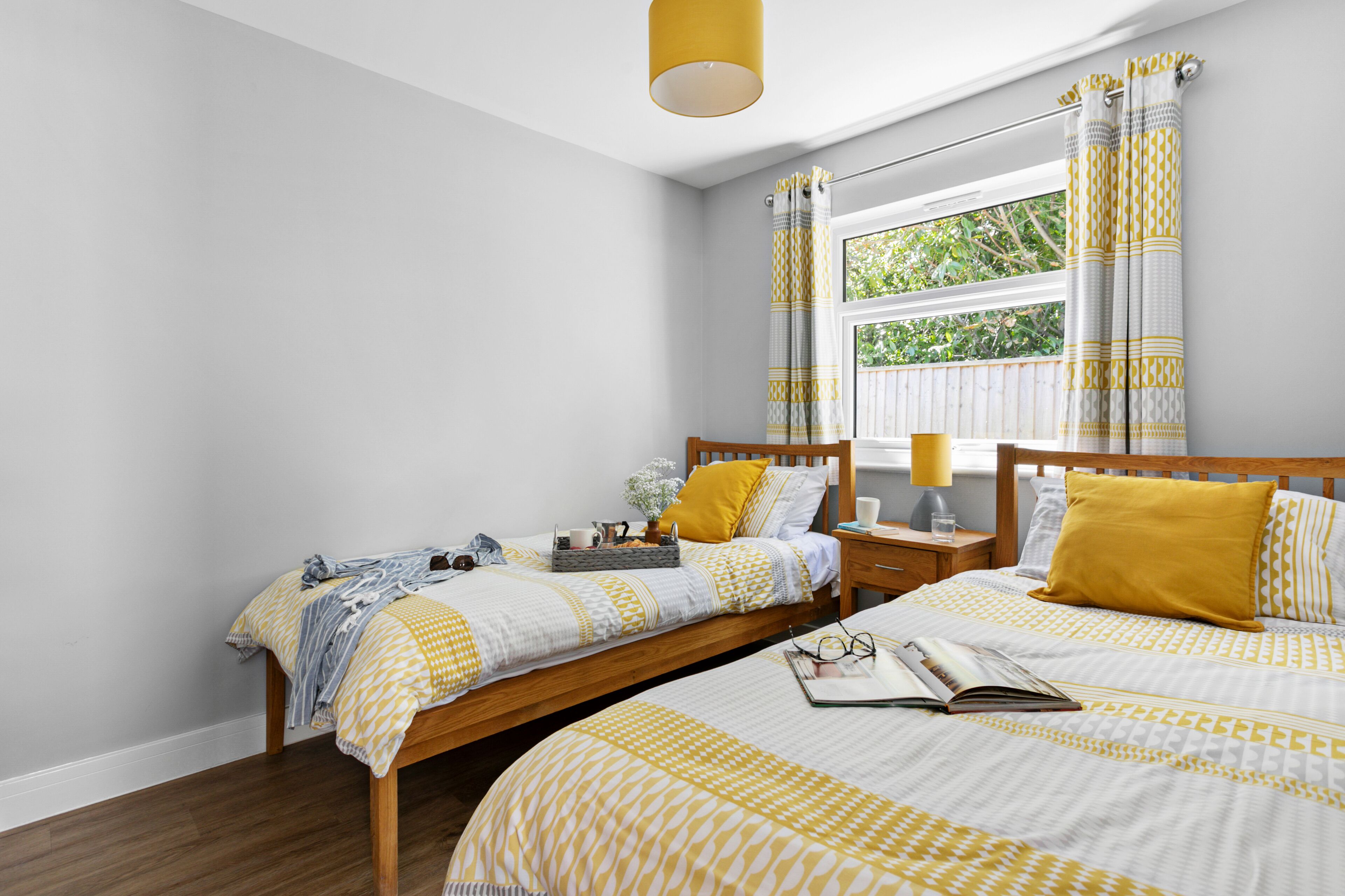 A bedroom with two single beds, a window with curtains, a bedside table with a lamp, and a nightstand with a lamp.