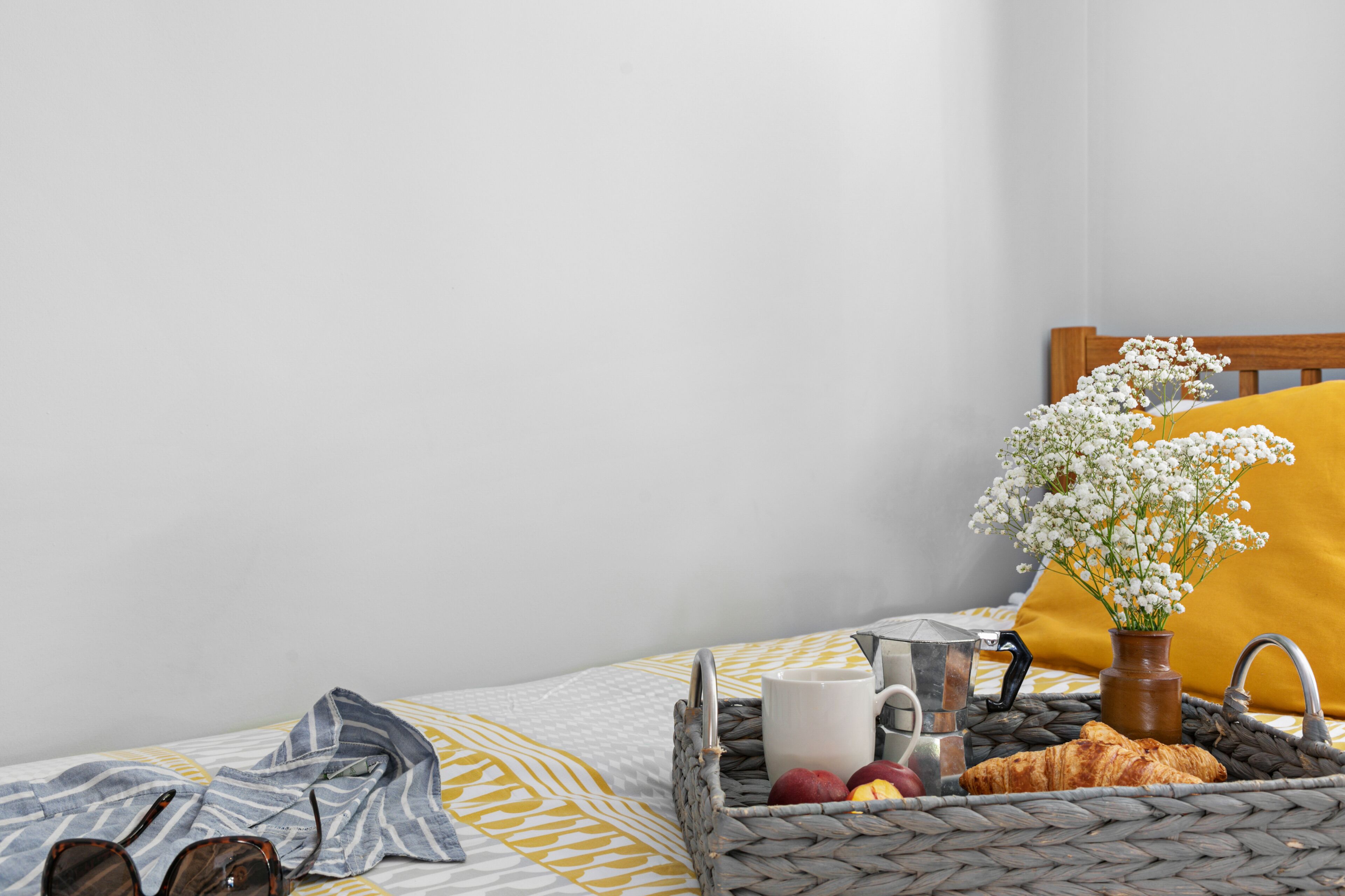 Ein ordentlich bezogenes Bett mit einer gestreiften Decke, ein Korbwäsche mit Croissants, eine Vase mit Blumen und eine Kaffeemaschine am Rand.