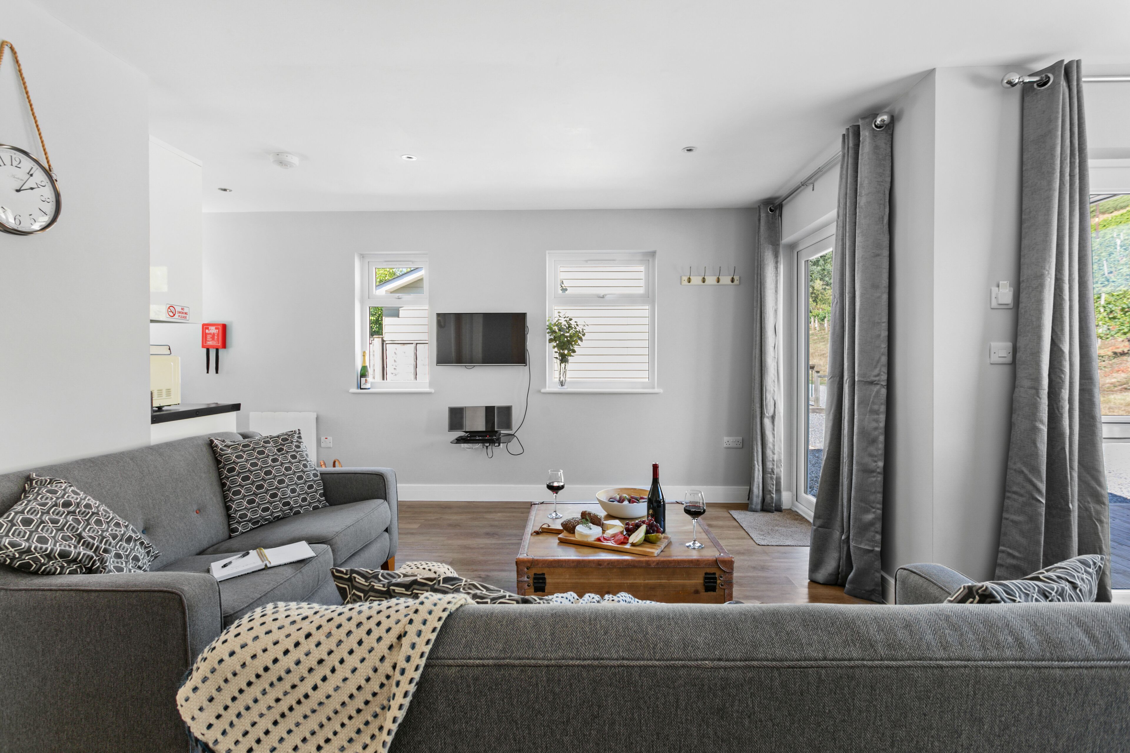Una sala de estar moderna con un sofá gris, una mesa de centro de madera con aperitivos y copas de vino, un televisor de pantalla plana y un ventanal con cortinas grises.