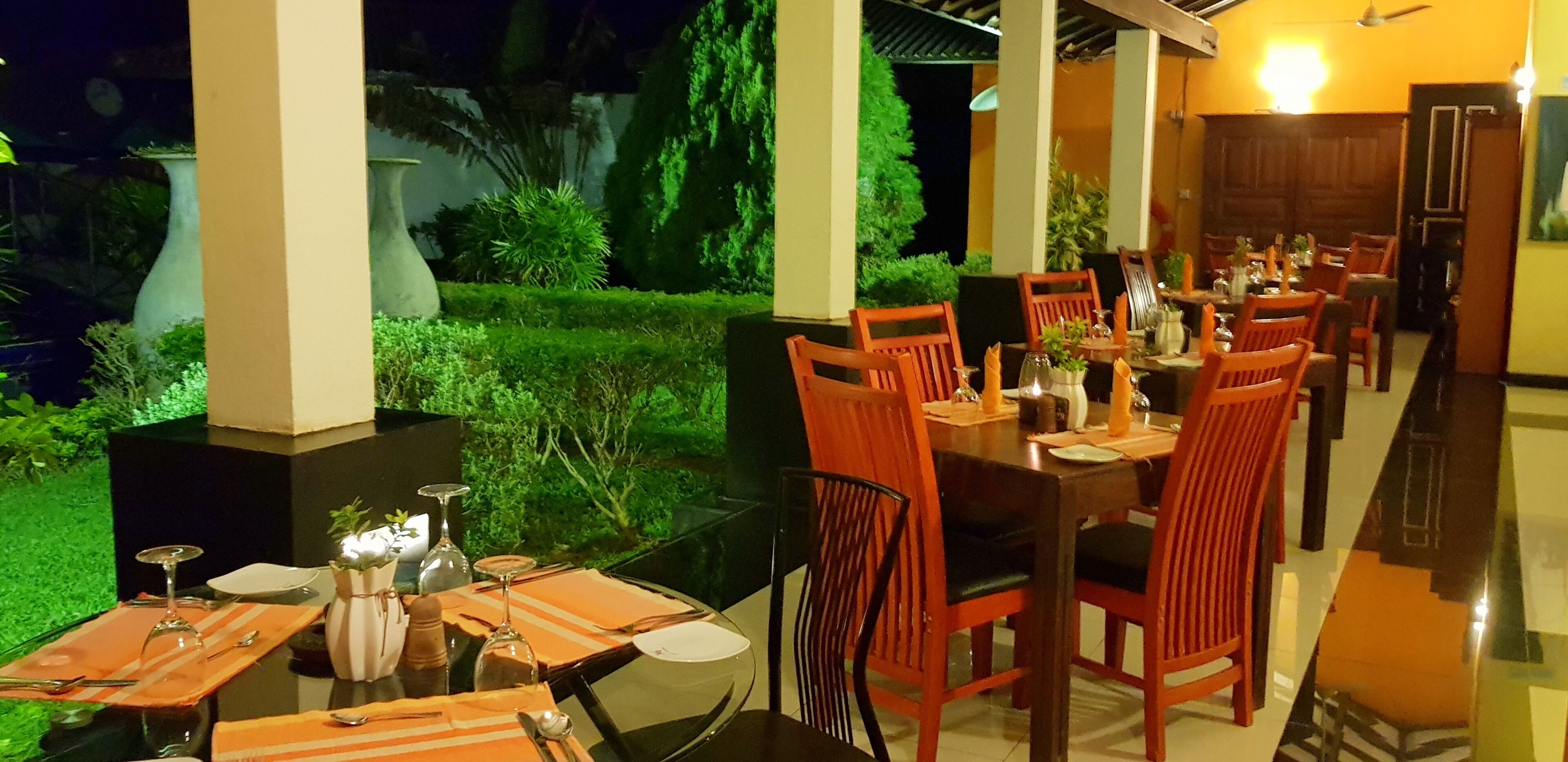 Uma sala de jantar com mesas preparadas para vários convidados, cercada por vegetação e vista para o céu noturno.