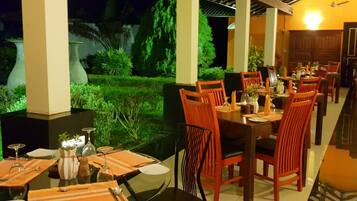 Uma sala de jantar com mesas preparadas para vários convidados, cercada por vegetação e vista para o céu noturno.