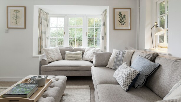 Ein gemütliches Wohnzimmer mit einem grauen Sofa, einem Holztisch mit einem Buch und einer Teekanne sowie einem Fenster mit Vorhängen.