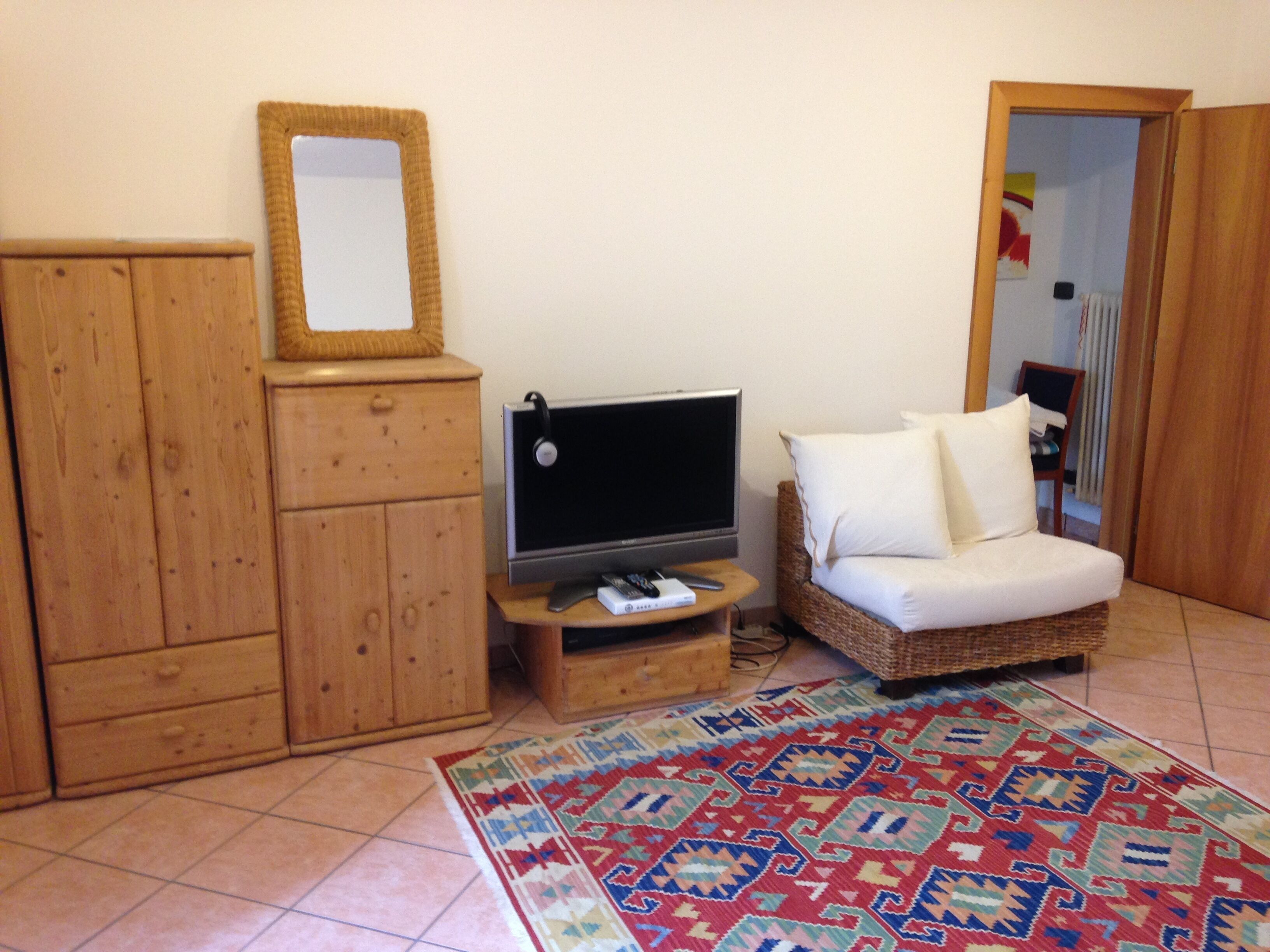 Ein Zimmer mit einem Holzschrank, einem Fernseher auf einem Holzgestell, einem Korbsessel mit weißen Kissen und einem gemusterten Teppich.