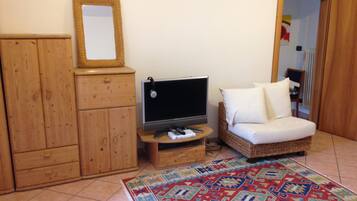 Ein Zimmer mit einem Holzschrank, einem Fernseher auf einem Holzgestell, einem Korbsessel mit weißen Kissen und einem gemusterten Teppich.