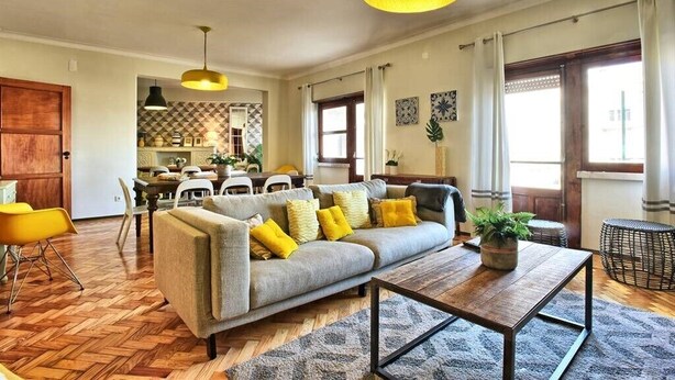 A spacious living room with a grey sofa, yellow cushions, a wooden coffee table, and a hanging light fixture.