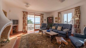 Una sala de estar con un sofá, sillones, una mesa de centro y un televisor. Se observa una escalera a la izquierda y vistas al mar a través de puertas de cristal.