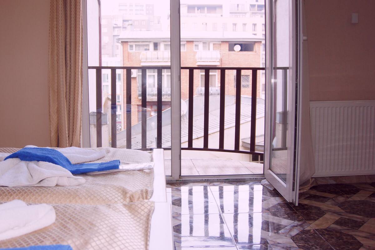 A hotel room with a bed, a window with a view of buildings, and a balcony with a railing.