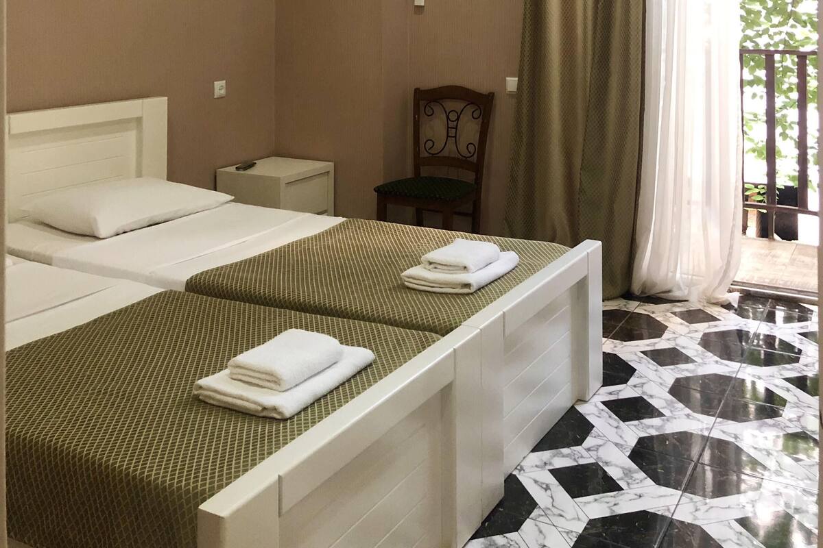 A hotel room with two beds, a chair, an air conditioning unit, and a view of greenery through the window.