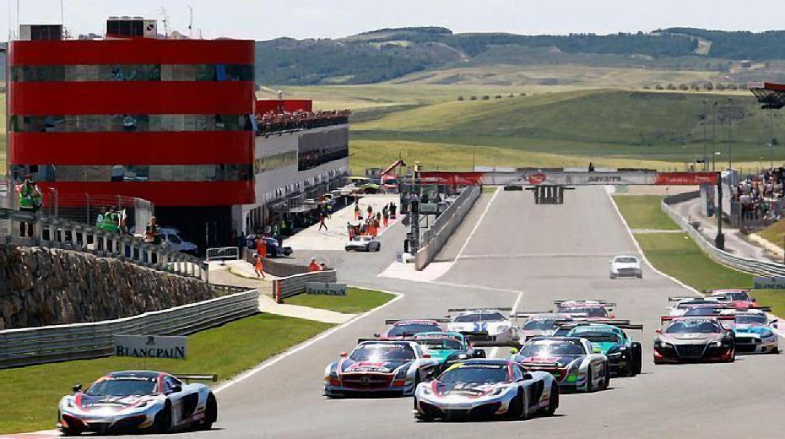 A race track with multiple racing cars lined up at the starting line, spectators, and a grandstand.
