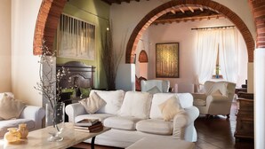 A cozy living room with a white sofa, a coffee table, and a fireplace. There are two armchairs and a painting on the wall. - Stunning private villa for 22 people with A/C, WIFI, private pool, TV, patio and panoramic view (Montebenichi)