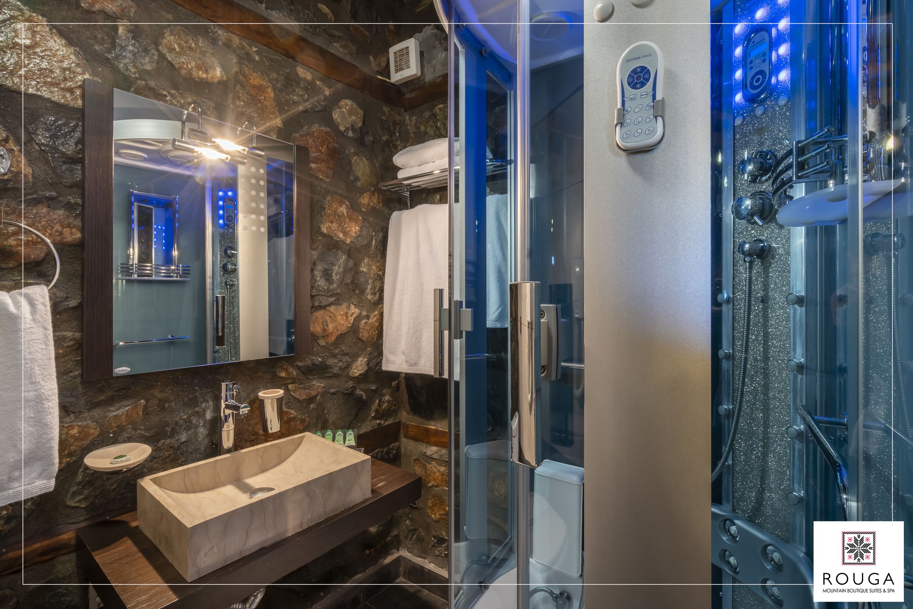 A modern bathroom with a stone wall, a wooden vanity, a large mirror, and a glass-enclosed shower.