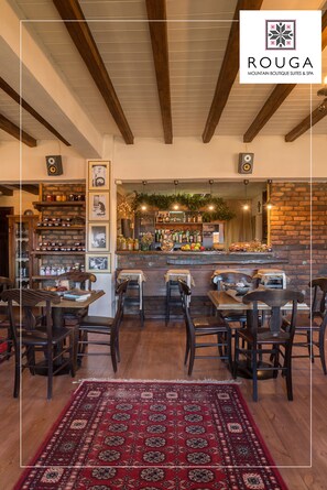 A cozy restaurant with wooden tables and chairs, a bar area with shelves of bottles, and a red patterned rug on the floor.