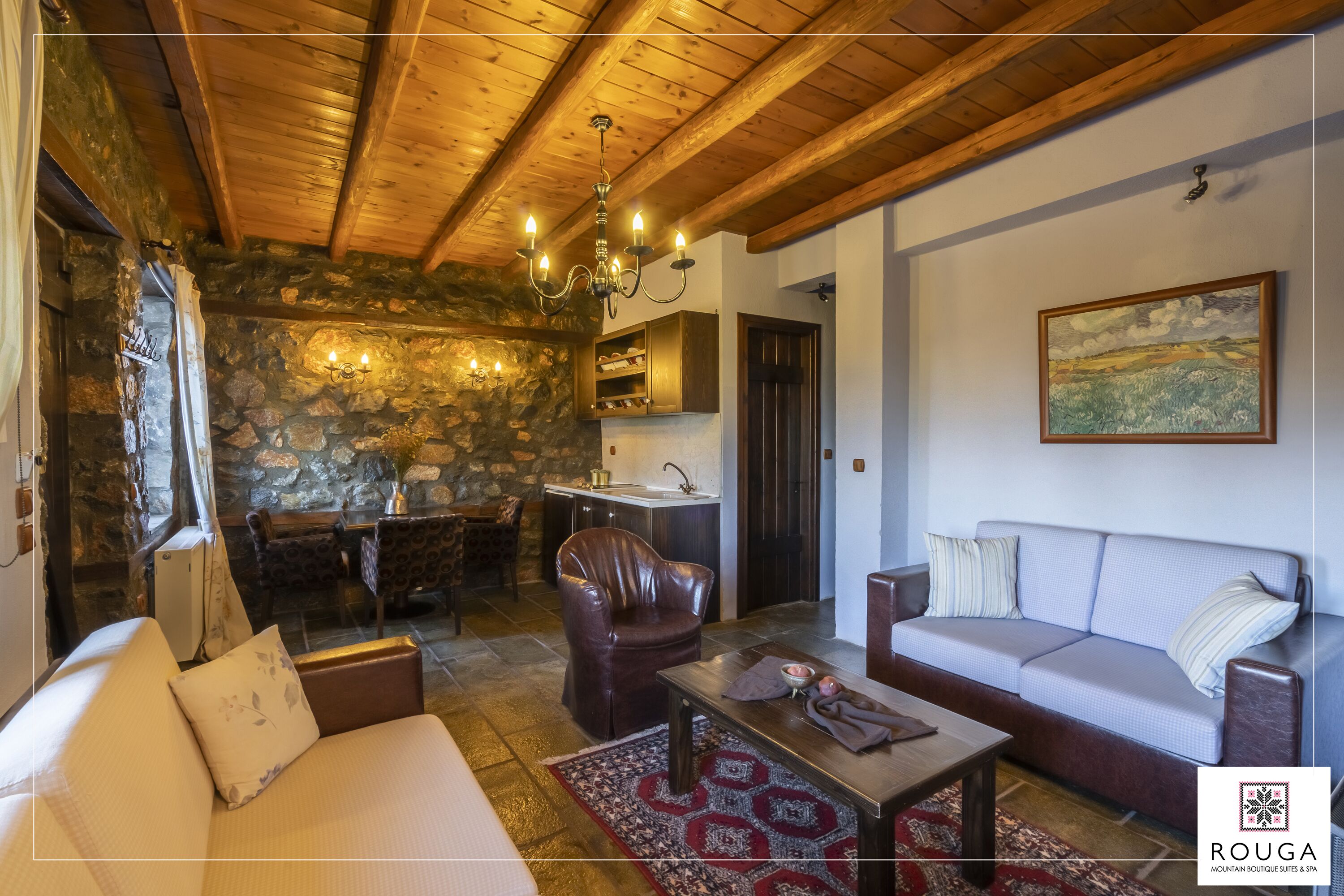 Un salon chaleureux avec un mur en pierre, des poutres apparentes en bois au plafond, un lustre, un canapé, un fauteuil en cuir et un coin repas agrémenté d’une cheminée en pierre.