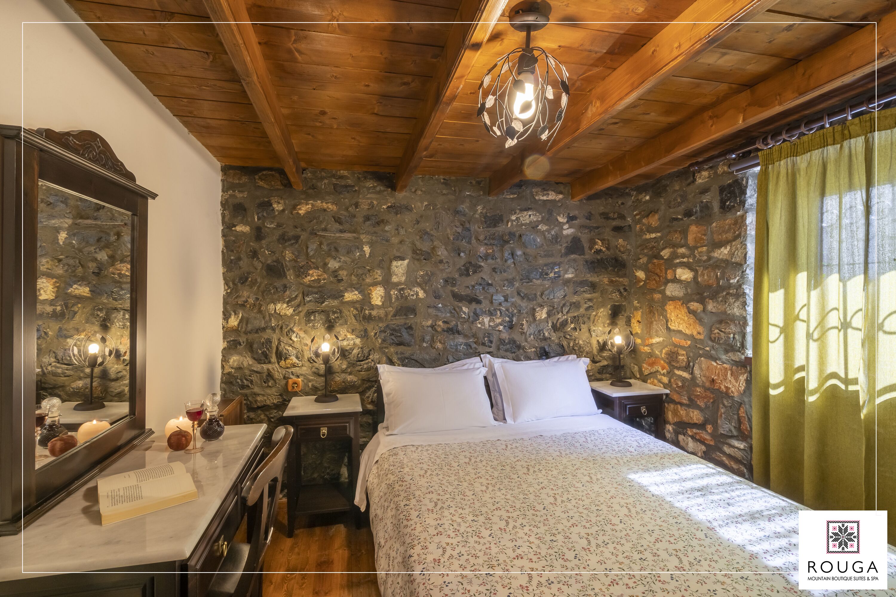 Quarto com parede de pedra, vigas de madeira no teto, cama com roupa de cama branca, luminárias de cabeceira e um espelho.